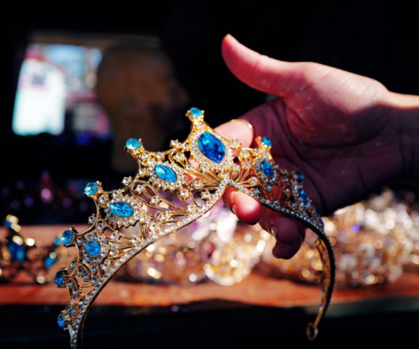 a hand holding a gold and diamond tiara with big blue gems in front of a table filled with crowns in different styles.