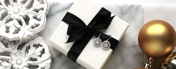 A marble tabletop with white rosette ornaments, a white box with a Christmas jewellery gift, wrapped with a black bow with two diamond earrings and gold Christmas balls.