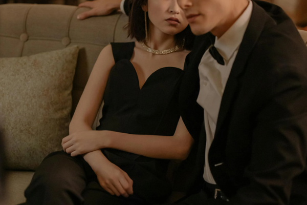 시크한 블랙 드레스와 눈부신 다이아몬드 목걸이, 테일러드 블랙 수트를 입은 젊은 커플이 소파에 우아하게 앉아 세련미와 고요한 사색에 잠긴 듯한 느낌을 자아냅니다.