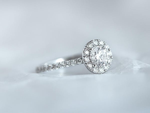 Il significato di un anello con halo - BAUNAT - Un raffinato anello di fidanzamento con castone a corona, che mette in risalto un diamante scintillante, adagiato su una superficie bianca immacolata.
