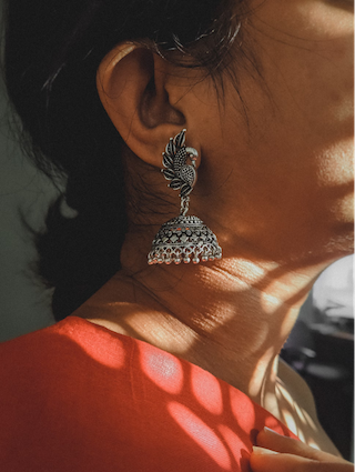 A woman in a vibrant red dress, adorned with earrings, showcasing cultural elegance with traditional jewellery of India - <strong>De unieke juwelenentraditie van India omvat met name tempeljuwelen, die worden gebruikt om goden en godinnen in tempels en heiligdommen te versieren. </strong>Deze juwelen bestaan meestal uit zware kettingen, ringen, oorbellen, neusringen en teenringen. Soortgelijke juwelen, die iets delicater zijn gemaakt, worden gedragen door de tempeldansers. Afhankelijk van de regio is het ontwerp van de juwelen strikt gebaseerd op bepaalde specificaties met betrekking tot de materialen, het aantal en type edelstenen, enz.
