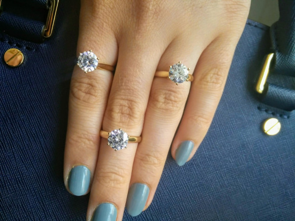 Woman's hand with blue manicure, wearing three rings including engagement ring with solitaire diamond gemstones - A selection of high-quality gold engagement rings with a solitaire from BAUNAT