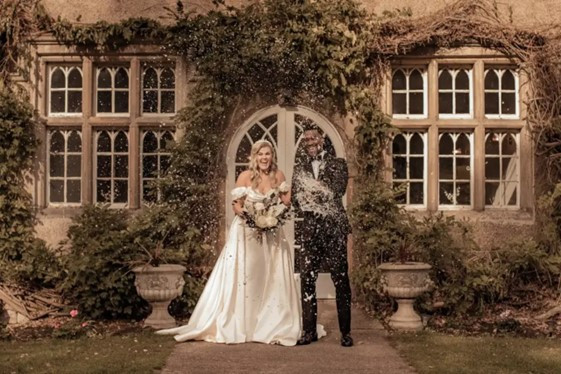 The bride and groom, dressed in classic English wedding attire, pose in front of a charming English wedding venue