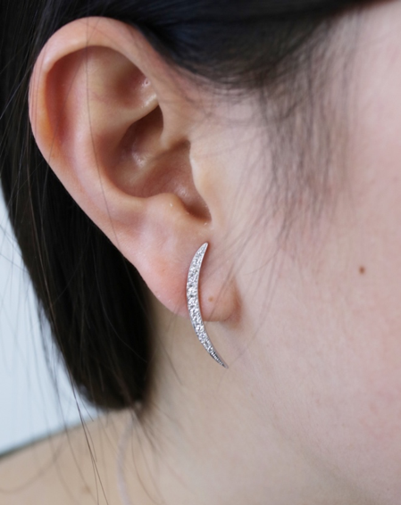 A close-up of a woman’s ear with a minimalistic white gold earring with diamonds. The earring is shaped like an elongated half-moon.
