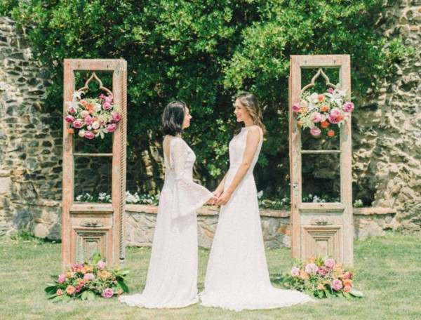 two brides smile in front of a wooden archway, surrounded by a beautiful outdoor setting during same-sex marriage in the UK
